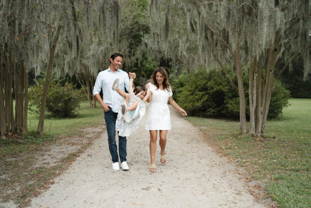 A man and woman walk along a tree-lined path, smiling as they swing a young girl between them by her arms. The surroundings are lush and green, with hanging moss on the trees.