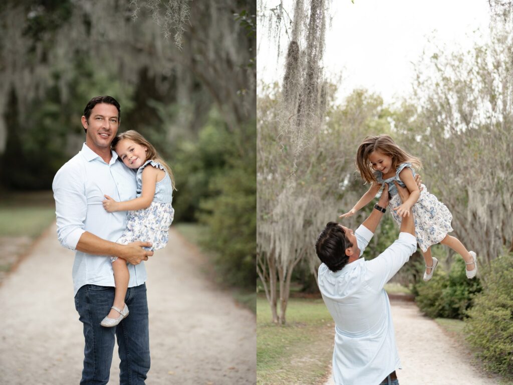 A man wearing a light blue shirt holds and plays with a young girl in a floral dress. They are outdoors on a tree-lined path, smiling and enjoying each other's company.