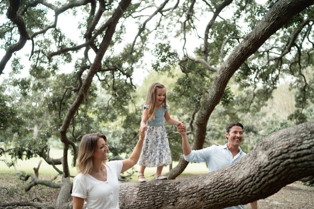 A smiling young girl balances on a large tree branch, holding hands with a woman and a man standing on either side, surrounded by leafy trees in a park setting.